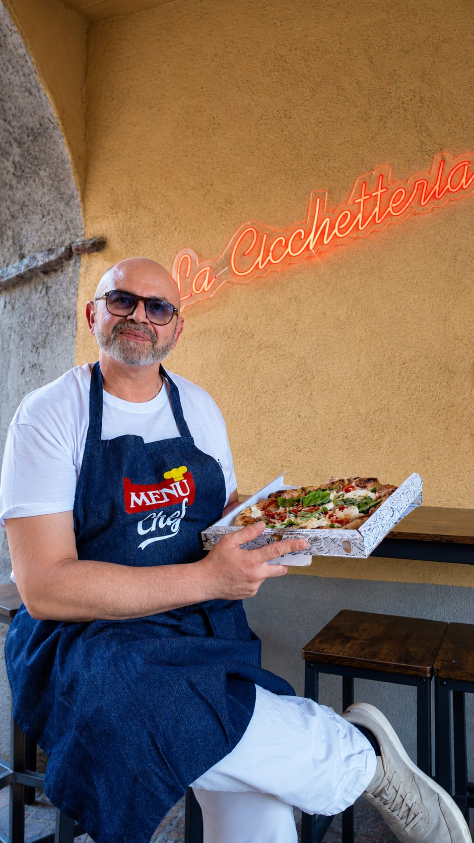 Enzo Ruocco con una pizza appena sfornata sotto l'insegna neon La Cicchetteria